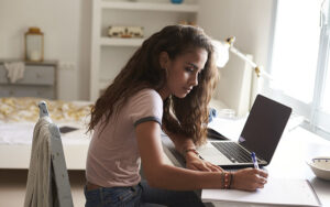 A young girl looking at her laptop and writing notes in a notebook at her desk in her room.