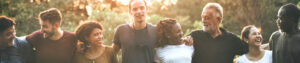 A group of people who are different races, ages, and genders standing shoulder to shoulder embracing one another outside in a park.