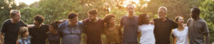 A group of people who are different races, ages, and genders standing shoulder to shoulder embracing one another outside in a park.