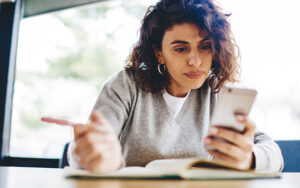 A young woman is looking at her phone, reading it, and taking notes.