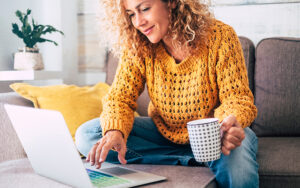 A middle-aged woman is holding a cup of coffee, reading her laptop, and sitting on her couch.