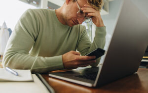 A young man is in distress and is holding his phone, while also looking at his laptop.