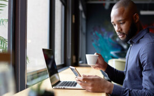 A young man at a coffee shop sipping coffee while working and looking at his phone.