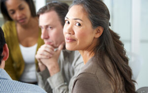 An older woman is sitting in a group of people, and looks back to the camera with a smile on her face.