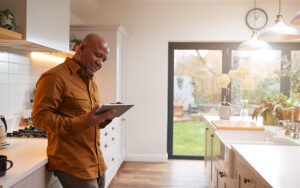 An older man is leaning against his kitchen cabinet reading his tablet.