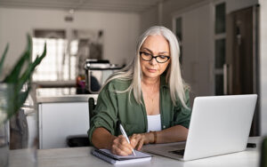 An elder woman taking notes on a pad of paper while sitting at her laptop.