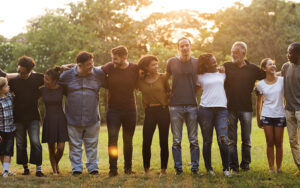 A group of people who are different races, ages, and genders standing shoulder to shoulder embracing one another outside in a park.