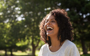 A young woman outside in a park laughing and smiling with the sun shining on her.