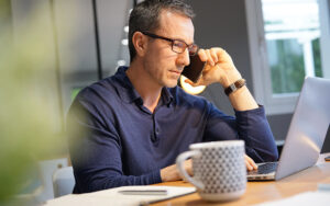 A man is talking on his cellphone while reading his computer.