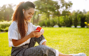 A young girl is sitting in the grass of a park listening to music and looking at her phone.