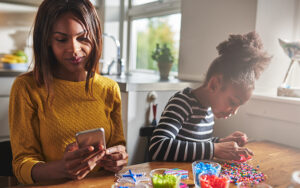 A mother and child are sitting at a kitchen table together. The mother is reading her phone while the child is making a craft.