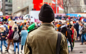 A person in a green coat and a black beanie hat looks out to a large group of people that are blurred in the photo.