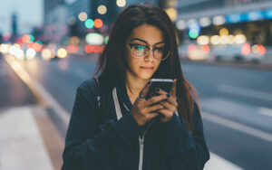 A woman is walking down a city block looking at her phone. The lights behind here are blurry and it's sunset.